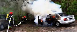 Na zdjęciu widzimy strażaków gaszących palący się samochód osobowy marki Audi. Strażacy ubrani w sprzęt ochrony układu oddechowego podają prąd piany na palący się samochód osobowy, z samochodu wydobywa się ogień i gęsty dym. W tle widać drzewa.