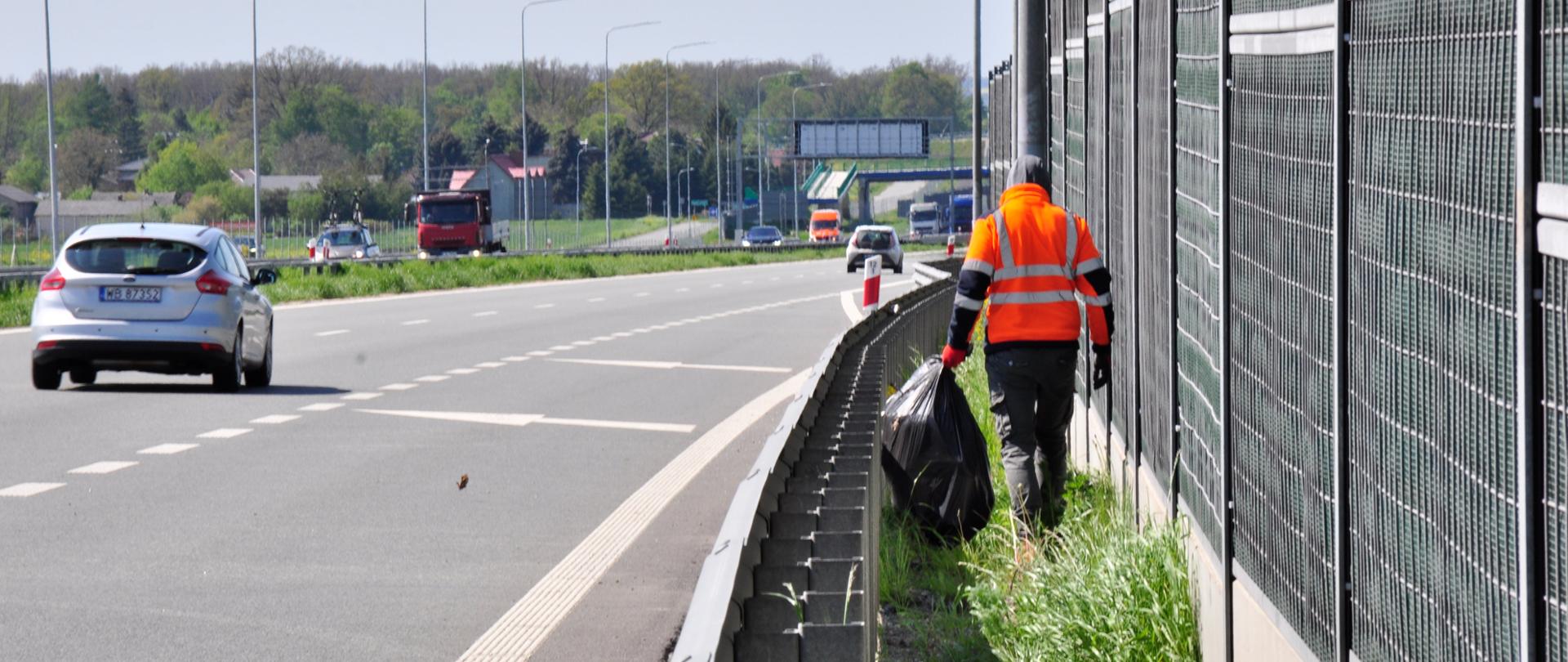 Zdjęcie przedstawia osobę ubraną w pomarańczową kamizelkę odblaskową, która porusza się wzdłuż pobocza autostrady, zbierając śmieci. Osoba ta trzyma duży czarny worek na śmieci i idzie obok metalowego ogrodzenia dźwiękochłonnego. Na drodze w tle widoczne są pojazdy, w tym srebrny samochód osobowy oraz czerwona ciężarówka. Po lewej stronie zdjęcia znajduje się pas zieleni oddzielający jezdnię od pobocza. W tle widoczne są drzewa oraz budynki, a dzień jest pogodny, z lekkim zachmurzeniem.