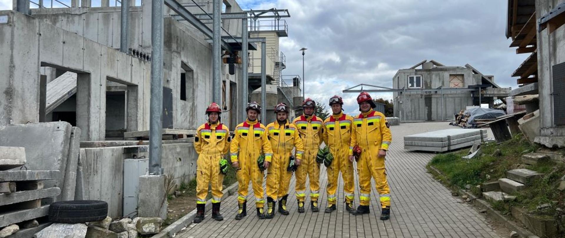 Zdjęcie przedstawia strażaków w trakcie szkolenia Specjalistycznej Grupy Poszukiwawczo-Ratowniczej. Ćwiczenia odbywają się na poligonie szkoleniowym w Gdańsku, w otoczeniu betonowych konstrukcji symulujących zawaliska. Strażacy w kombinezonach ochronnych i kaskach wykonują zadania z zakresu przeszukiwania gruzowisk oraz zabezpieczania konstrukcji. Widać instruktora nadzorującego przebieg zajęć. Fotografia dokumentuje praktyczne szkolenie w realistycznych warunkach terenowych.