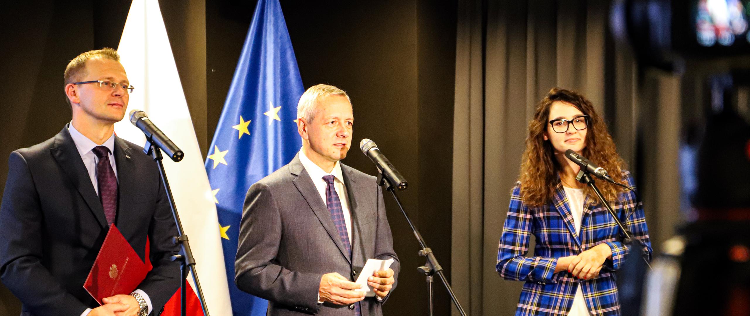 Konferencja prasowa. Trzy osoby stoją przed mikrofonami. Od lewej: Hubert Nowak, Marek Zagórski i Justyna Orłowska. W tle flagi: polska i unijna