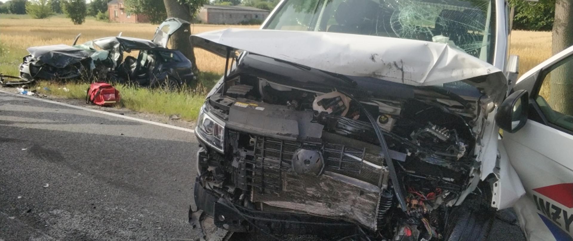Na zdjęciu samochody uczestniczące w zdarzeniu - na pierwszym planie stojący na jezdni samochód dostawczy koloru białego z rozbitym przodem. Na drugim planie wrak samochodu osobowego koloru ciemnego.