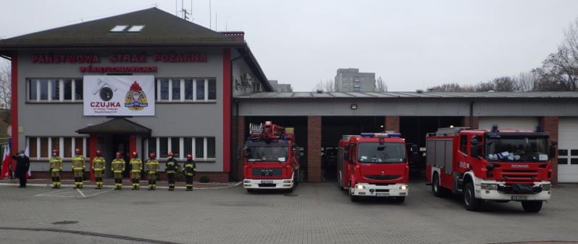Zdjęcie przedstawia strażaków stojących na baczność przed budynkiem komendy miejskiej Państwowej Straży Pożarnej w Świętochłowicach. Po prawej stronie przed bramami garażowymi stoją zaparkowane samochody pożarnicze.