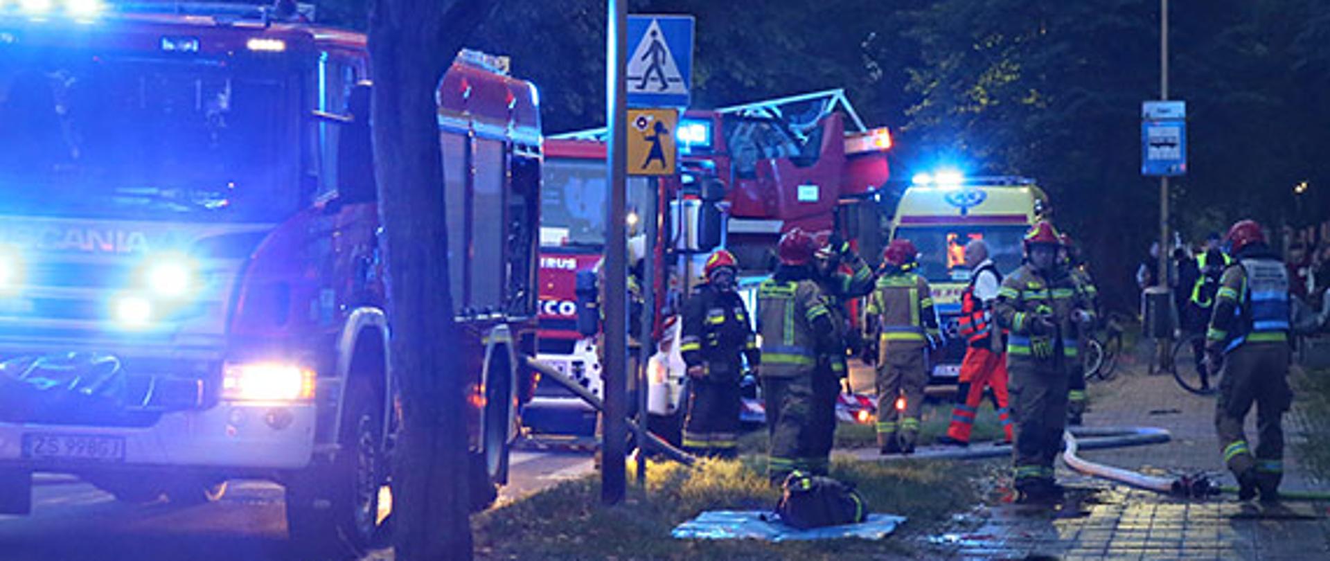 Zdjęcie przedstawia strażaków w trakcie działań. Po lewej stronie widoczne samochody pożarnicze.