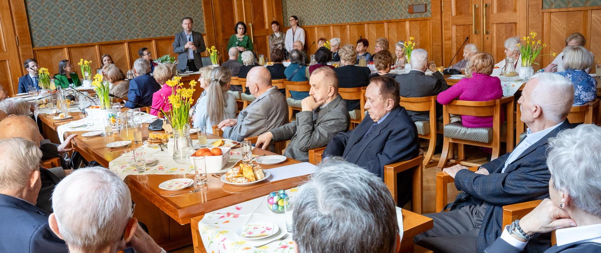 Zdjęcie z boku, przy stołach ustawionych w literę U siedzą ludzie, na stole dekoracja wielkanocna. Mężczyzna w garniturze stoi i przemawia. 