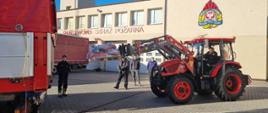Strażacy za pomocą traktora z widłami ładują palety ze sprzętem na naczepę samochodu.