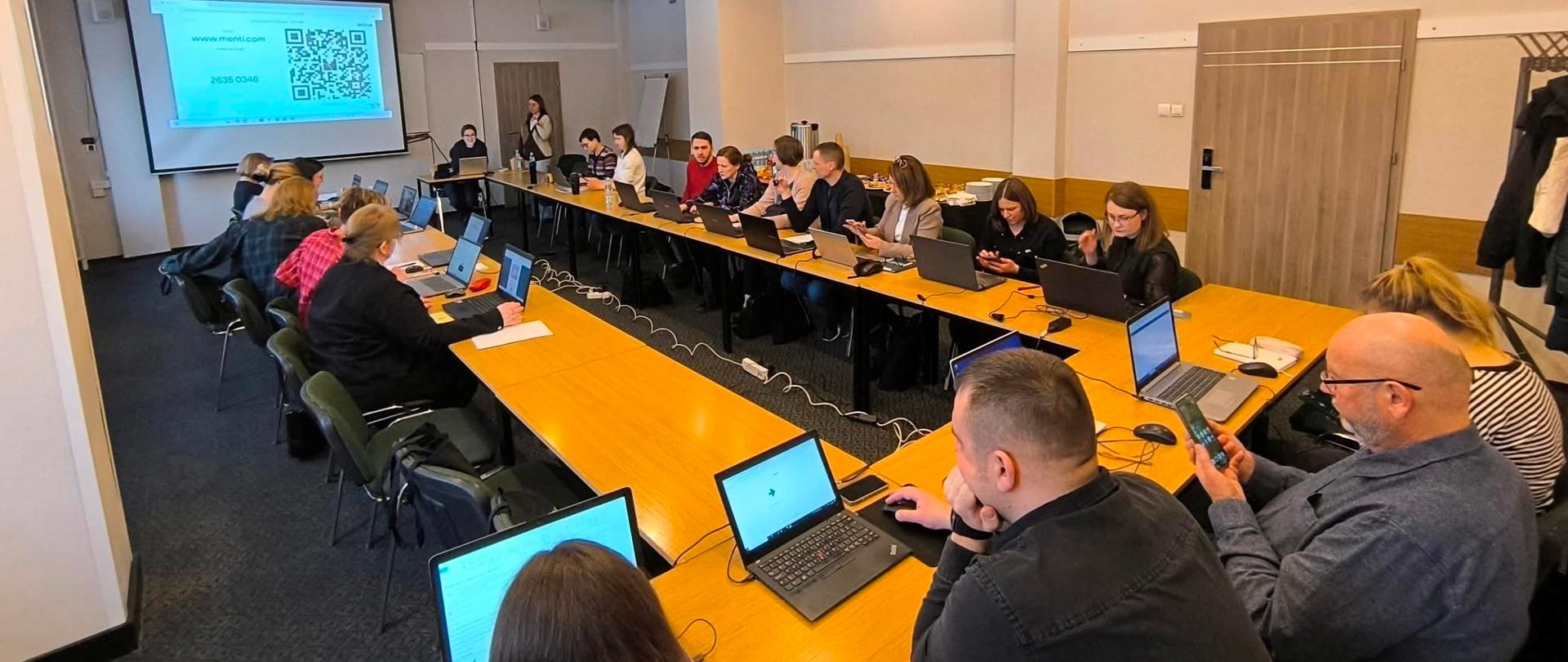 Na zdjęciu widać jasną, nowoczesną salę szkoleniową lub konferencyjną, w której odbywa się spotkanie warsztatowe. Ustawione w kształt litery U drewniane stoły wypełniają niemal całe pomieszczenie. Przy stołach siedzi kilkanaście osób – kobiety i mężczyźni w różnym wieku – większość pracuje na otwartych laptopach. Część uczestników patrzy w ekrany komputerów, inni trzymają w rękach telefony komórkowe. Na blatach leżą notatniki, myszki komputerowe i przewody zasilające; wzdłuż stołów poprowadzone są przedłużacze z podłączonymi ładowarkami.
Na końcu sali znajduje się ekran projekcyjny. Wyświetlona jest na nim strona internetowa menti.com z dużym kodem QR oraz krótkim kodem liczbowym do dołączenia do interaktywnej ankiety. Obok ekranu stoi osoba prowadząca – kobieta trzymająca mikrofon – a przy stole obok siedzi druga osoba obsługująca komputer.
Pomieszczenie ma jasne ściany i sufit podwieszany z równomiernie rozmieszczonymi punktowymi lampami. W suficie widoczna jest klimatyzacja kasetonowa. Po prawej stronie znajduje się drewniane drzwi oraz wieszak z powieszonymi płaszczami i kurtkami. Atmosfera wygląda na skupioną i roboczą – uczestnicy są zaangażowani w ćwiczenie lub ankietę online podczas szkolenia.