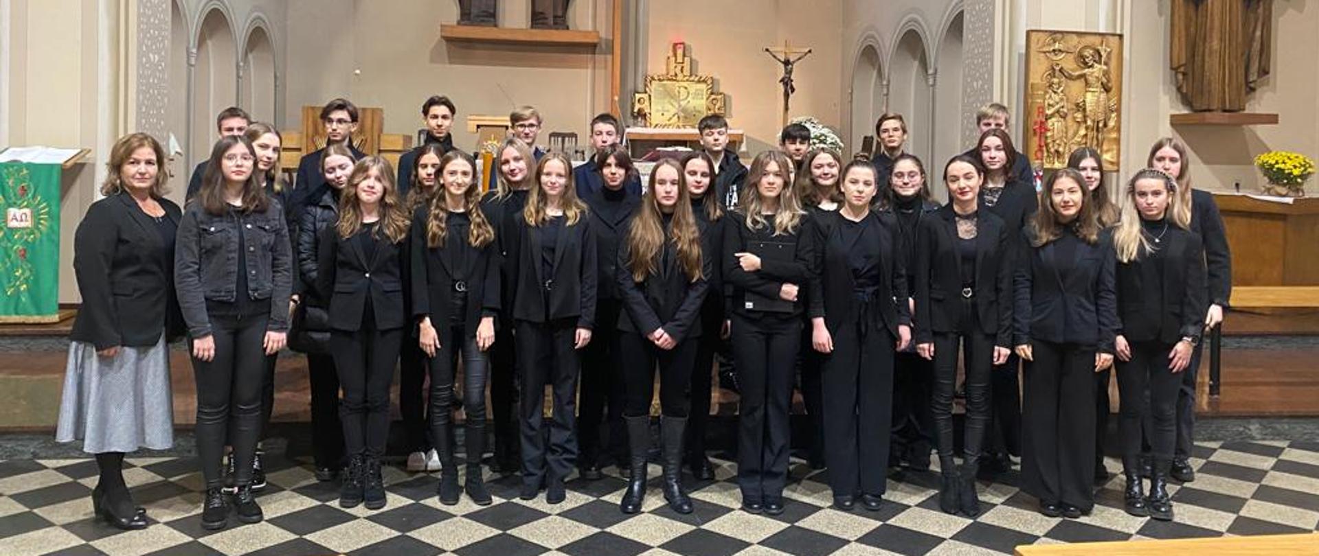 Kolorowe zdjęcie chóru Dominanta. Chór ustawiony jest w trzech rzędach (dwa pierwsze to dziewczyny, trzeci to chłopacy) na schodach przed ołtarzem w kościele p.w. Św. Józefa Robotnika. Wszyscy członkowie ubrani są w czarne stroje, poza panią dyrygent, która ma srebrną spódnicę.