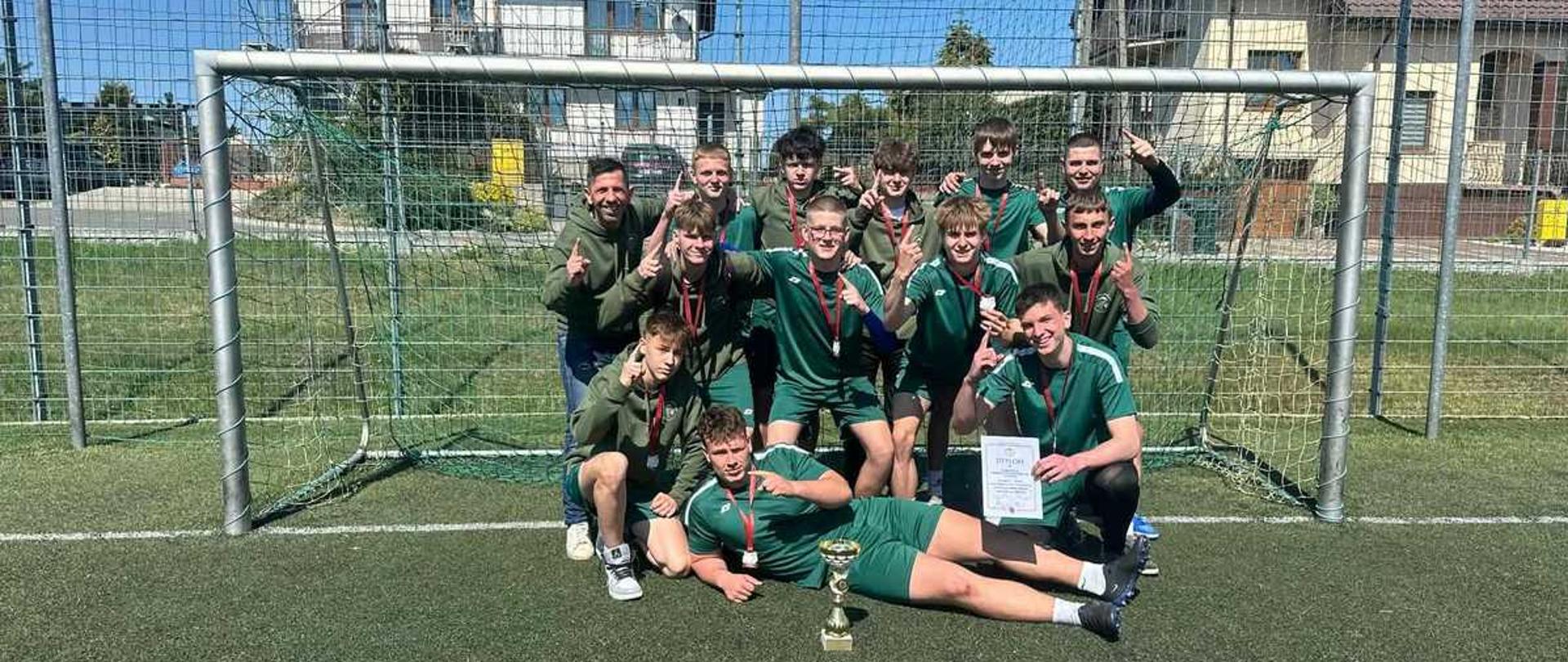Zdjęcie przedstawia grupę dwunastu młodych mężczyzn, prawdopodobnie drużynę piłkarską, świętującą sukces na boisku ze sztuczną nawierzchnią.
Kompozycja i sceneria
Mężczyźni pozują na tle bramki piłkarskiej. Dzień jest słoneczny, a niebo bezchmurne. W tle widać domy jednorodzinne oraz metalowe ogrodzenie boiska.
Osoby na zdjęciu
Większość zawodników ubrana jest w pasujące do siebie, ciemnozielone stroje sportowe (koszulki i krótkie spodenki). Niektórzy mają na sobie bluzy z kapturem w tym samym kolorze. Na szyjach wielu z nich wiszą medale na czerwonych tasiemkach.
Pierwszy plan: Dwóch młodych ludzi leży lub półleży na murawie przed resztą grupy. Jeden z nich, po prawej, trzyma w dłoni biały dyplom. Pomiędzy nimi, bezpośrednio na trawie, stoi złoty puchar.
Środkowy plan: Grupa osób klęczy lub kuca.
Ostatni plan: Pozostali zawodnicy stoją wewnątrz bramki lub tuż przed nią.
Atmosfera i detale
Wszyscy są uśmiechnięci i wyraźnie cieszą się z wygranej. Wielu z nich wykonuje gest uniesionego palca wskazującego (symbolizujący „numer jeden”). Po lewej stronie grupy stoi nieco starszy mężczyzna, prawdopodobnie trener, ubrany w zieloną bluzę i jeansy.