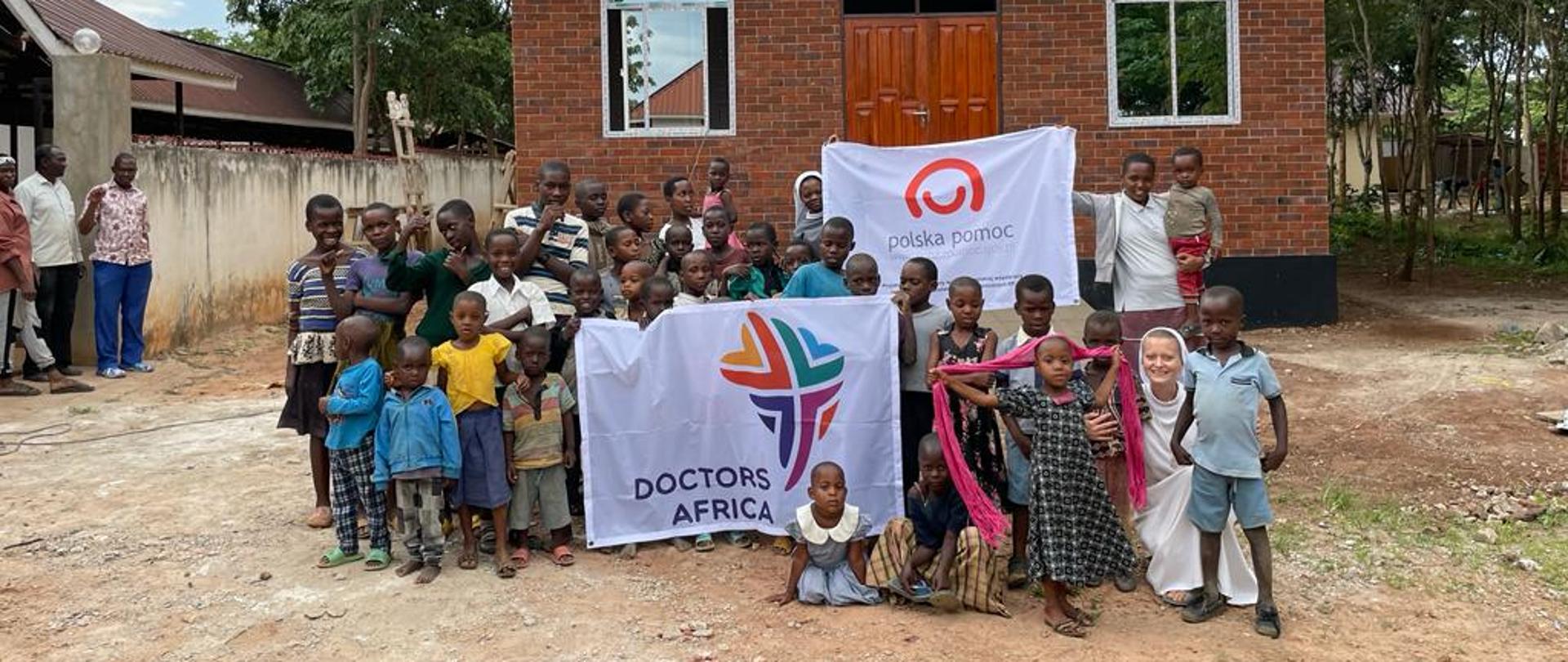 Grupa kilkudziesięciu osób (głównie dzieci) pozuje do zdjęcia przed nowowybudowanym parterowym budynkiem. Trzymają przed sobą banery z logotypami Doctors Africa i polska pomoc.