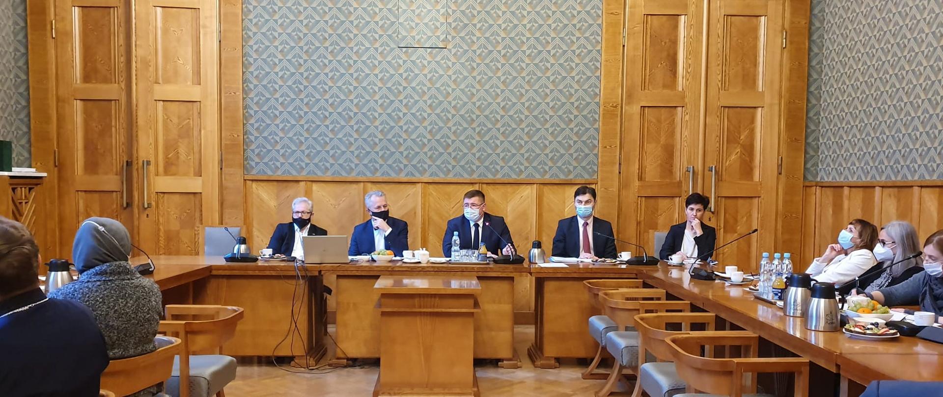 Duża sala z drewnianą boazerią. Wokół stołu ustawionego w podkowę siedzą przedstawiciele kościołów i związków wyznaniowych. Przy stole prezydialnym wiceminister Tomasz Rzymkowski i pracownicy Ministerstwa Edukacji i Nauki. 