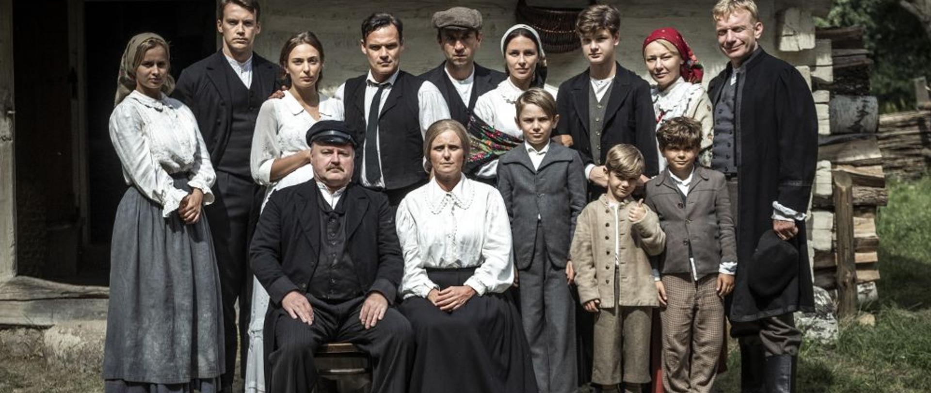 Przed wiejską chatą siedzą dziadkowie, a za ich plecami stoją ich dzieci i wnuki.