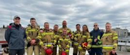 Grupa strażaków PSP i SFRS w ubraniach specjalnych pozuje na terenie poligonu; w rękach hełmy, w tle zabudowa miejska i pochmurne niebo.