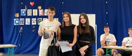 "Na zdjęciu widać uroczystość szkolną z okazji „Dnia Chłopaka”, która odbywa się w hali sportowej lub sali gimnastycznej.
Oto szczegółowy opis elementów znajdujących się na fotografii:
• Pierwszy plan (środek): Na środku sali stoją trzy nastoletnie osoby – jeden chłopak i dwie dziewczyny.
• Chłopak (po lewej): Ma krótkie, ciemne włosy, ubrany jest w jasny t-shirt z nadrukiem, jasne, luźne spodnie bojówki oraz ciemne buty sportowe. W jednej ręce dumnie trzyma pamiątkowy dyplom lub certyfikat, a na ramieniu ma przewieszoną czarną, materiałową torbę. Lekko się uśmiecha.
• Dziewczyna w środku: Ma proste, brązowe włosy sięgające ramion. Jest ubrana w czarną bluzkę bez rękawów, szerokie czarne spodnie z paskiem i ciemne buty sportowe. W rękach trzyma plik kartek z dokumentami.
• Dziewczyna po prawej: Ma długie, ciemne włosy, ubrana jest w czarną bluzkę z długim rękawem, czarne spodnie oraz białe buty sportowe. W dłoni trzyma mikrofon, co sugeruje, że prowadzi uroczystość.
• Drugi plan (tło):
• Za stojącymi osobami zawieszona jest wielka, ciemnoniebieska kurtyna, która zasłania ścianę. Na kurtynie po lewej stronie widnieje duży, kolorowy napis wykonany z ozdobnych liter: „DZIEŃ CHŁOPAKA”. Dekorację uzupełniają drobne elementy, takie jak czerwone serce i mała papierowa piłka nożna.
• Po prawej stronie, w głębi, przy stolikach sędziowskich lub szkolnych, siedzą dwaj inni chłopcy w białych t-shirtach i krótkich spodenkach, którzy przyglądają się wydarzeniu.
• Konstrukcja sali (góra i dół):
• Sufit: Nad całą sceną unosi się wysoki, łukowaty sufit hali o charakterystycznej konstrukcji z drewnianych kantówek i metalowych rur wentylacyjnych. W centralnym punkcie pod sufitem wisi złożona konstrukcja z tablicą i obręczą do koszykówki. Od sufitu ciągnie się także ochronna siatka siatkarska.
• Podłoga: Całość odbywa się na szarej, gładkiej nawierzchni sportowej."