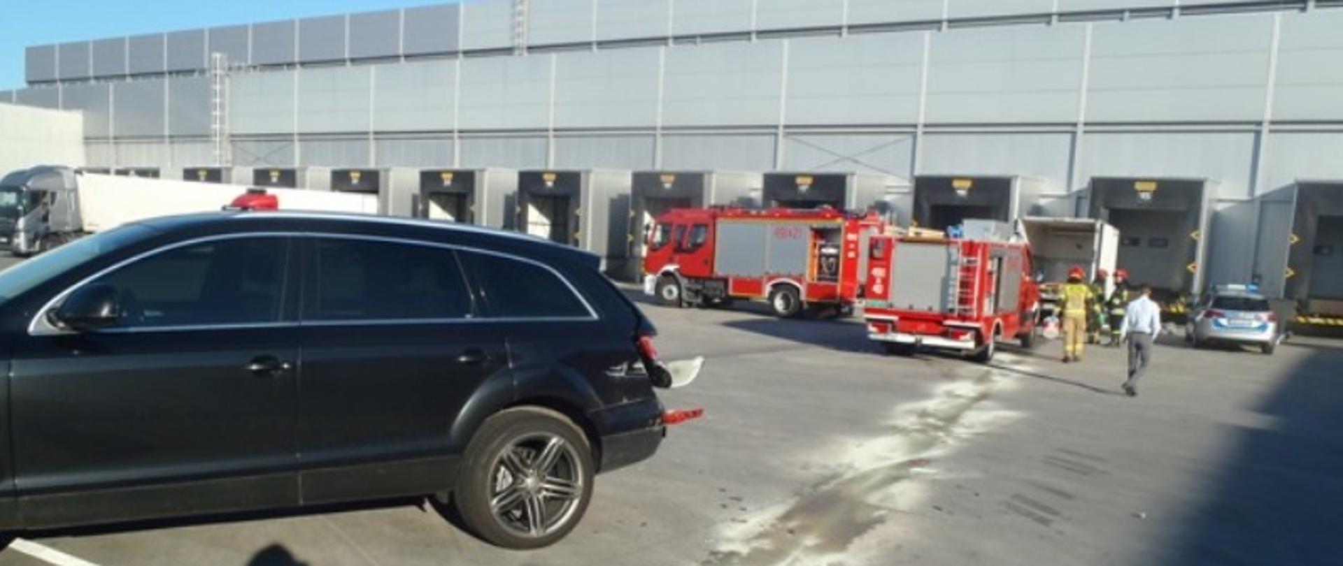 Na pierwszym planie czarny duży samochód osobowy marki Audi z uszkodzonym lewym bokiem. Za uszkodzonym pojazdem w odległości około pięciu metrów stoją dwa pojazdy pożarnicze, radiowóz oraz samochód dostawczy z otwartą skrzynią ładunkową. Samochód dostawczy przodem oparty jest o rampę załadunkową dużego magazynu. Obok samochodu dostawczego znajdują się strażacy, którzy zabezpieczają miejsce zdarzenia. Na placu załadunkowym widać plamę oleju, która została zabezpieczona sorbentem,. W kierunku miejsca prowadzonych działań zmierza osoba cywilna w koszuli, prawdopodobnie kierownik magazynu. 