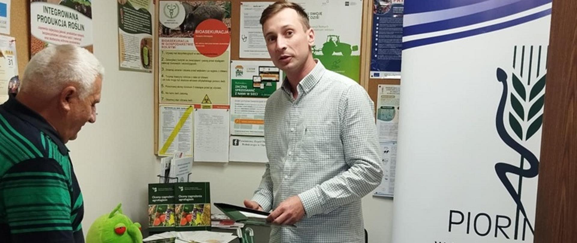 Stoisko w postaci czarnego stolika z krzesłem, wokół na ścianie znajdują się różnokolorowe ulotki dotyczące zagadnień rolniczych. Na pierwszym planie widać mężczyznę ubranego w jasną koszulę oraz niebieskie spodnie- pracownika inspekcji. Widać również mężczyznę niskiego wzrostu w czarno zielonej koszulce, któremu pracownik tłumaczy interesujące go zagadnienie zawarte w ulotce. Na stoliku leży zielona maskotka z długim nosem oraz czerwonymi oczami będąca maskotką kampanii ,, Dbaj o zdrowie, chroń życie’’. Po prawej stronie widać biało granatowy roll-up z logo inspekcji