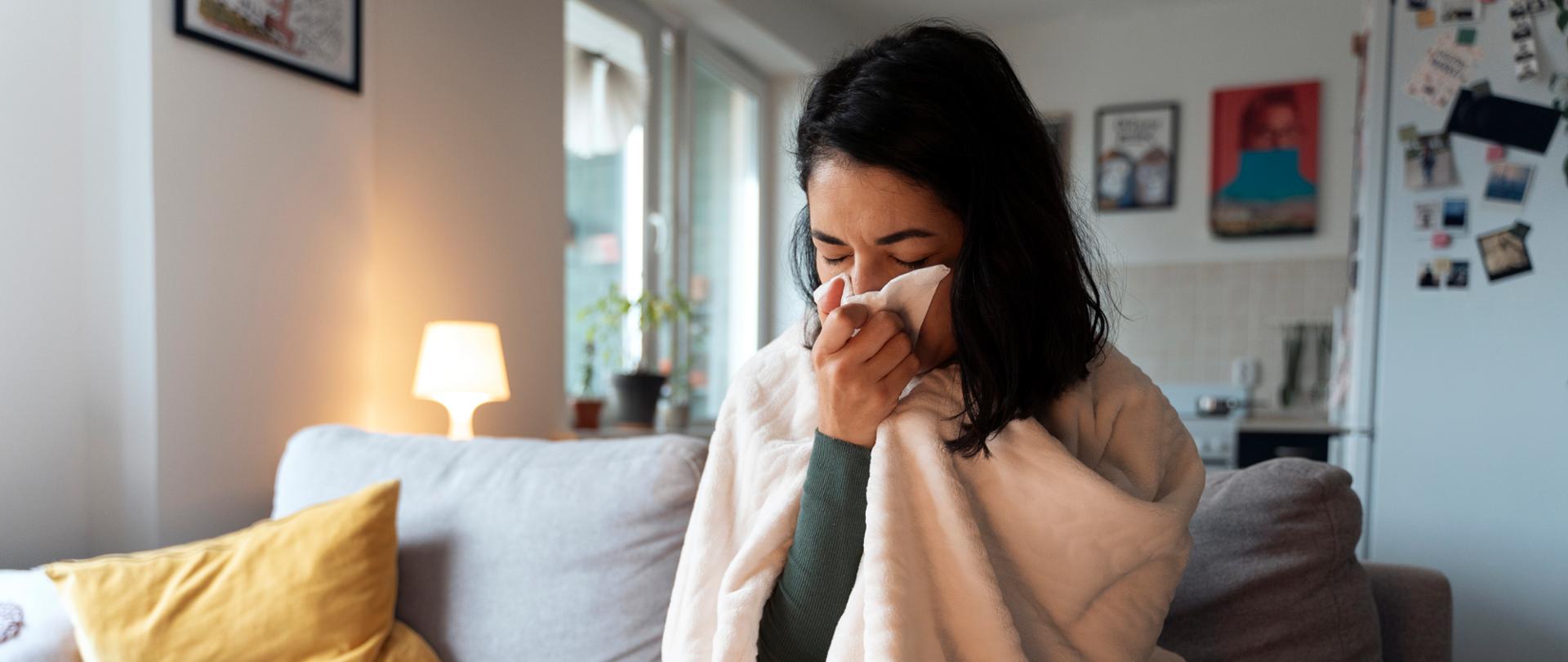 Zdjęcie przedstawia kobietę chorą na grypę, która leczy się w domu. Jest owinięta kocem oraz ma katar.
