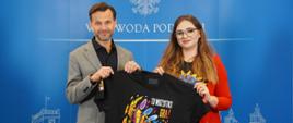 Wojewoda podlaski Jacek Brzozowski wspólnie z Natalią Stieszenko trzymają czarną koszulkę z grafiką 32 finału WOŚP