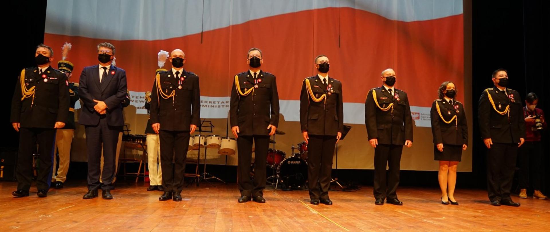 Sekretarz Stanu w MSWiA oraz MWK PSP stoją obok 6 odznaczonych funkcjonariuszy na scenie na tle biało - czerwonej flagi.