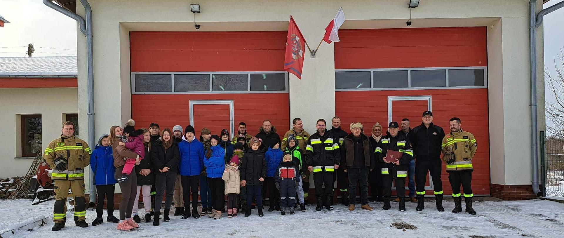 Na tle remizy OSP z dużymi czerwonymi drzwiami stoi grupa strażaków w mundurach oraz dzieci. Zima, biało od śniegu.
