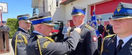 Zdjęcie przedstawia teren Jednostki Ratowniczo-Gaśniczej oraz strażaków ubranych w mundury galowe koloru czarnego ze sznurem galowym oraz czapką rogatywką na głowie, podczas obchodów Dnia Strażaka w Radziejowie. Na zdjęciu Kujawsko-Pomorski Komendant Wojewódzki Państwowej Straży Pożarnej w asyście Komendanta Powiatowego Państwowej Straży Pożarnej wręcza odznaczenia