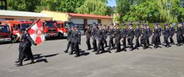 Maszerują strażacy na początku poczet sztandarowy a za nim pododdział w mundurach wyjściowych. W tle widać samochody pożarnicze oraz budynki strażackie i drzewa.