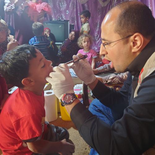 Lekarz bada gardło chłopczyka; w tle grupa kobiet i dzieci czeka na badanie