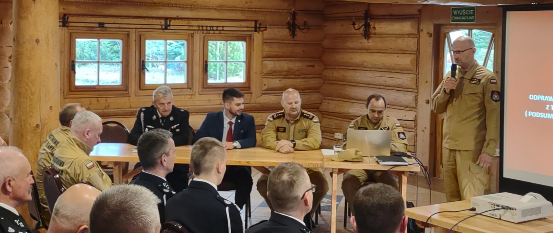 Komendant powiatowy PSP w Żyrardowie st. bryg. Mariusz Tymoszewicz przemawia do mikrofonu stojąc przodem do ochotników. Po jego prawej stronie siedzą kolejno mł. asp. Tomasz Badowski, zastępca mazowieckiego komendanta PSP st. bryg. Tomasz Cybul, starosta powiatu żyrardowskiego Krzysztof Dziwisz, dh Waldemar Suski.