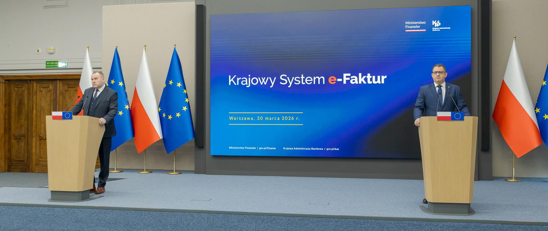 Marcin Łoboda szef Krajowej Administracji Skarbowej oraz Zbigniew Stawicki zastępca szefa KAS podczas konferencji prasowej w Ministerstwie Finansów opowiedzieli o wdrożeniu II etapu Krajowego Systemu e-Faktur przy mównicach przed dużym ekranem z napisem „Krajowy System e‑Faktur”, obok flag Polski i Unii Europejskiej