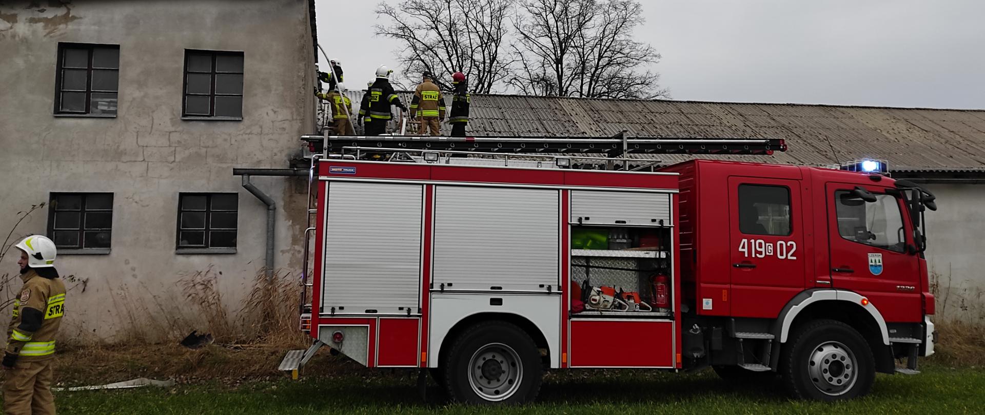 Wóz strażacki widziany z boku przed budynkiem, na dachu tego budynku strażacy prowadzą działania. 