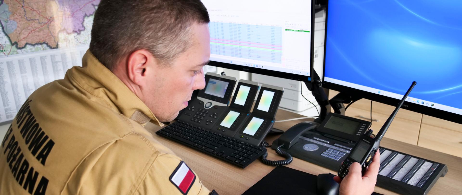 Strażak w mundurze musztardowym siedzi przy stole na którym leżą radiotelefony oraz są przymocowane monitory funkcjonariusz w ręce trzyma radiotelefon.