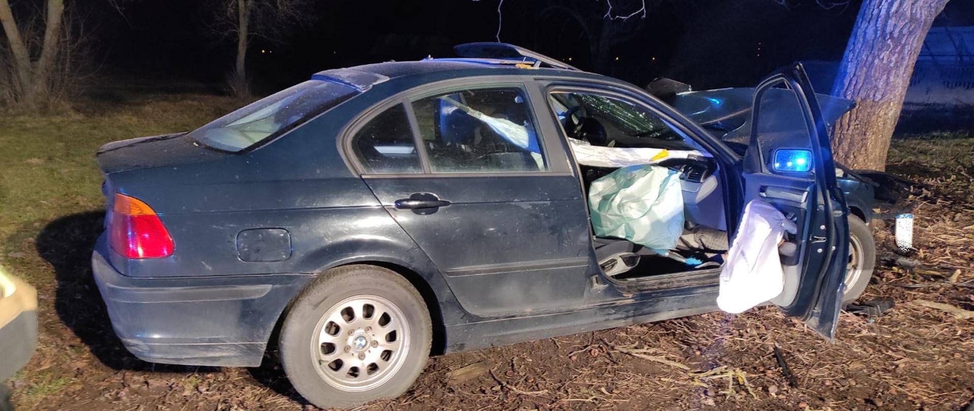 Rozbity samochód osobowy ciemnego koloru. Otwarte przednie drzwi. W środku otwarte poduszki powietrzne