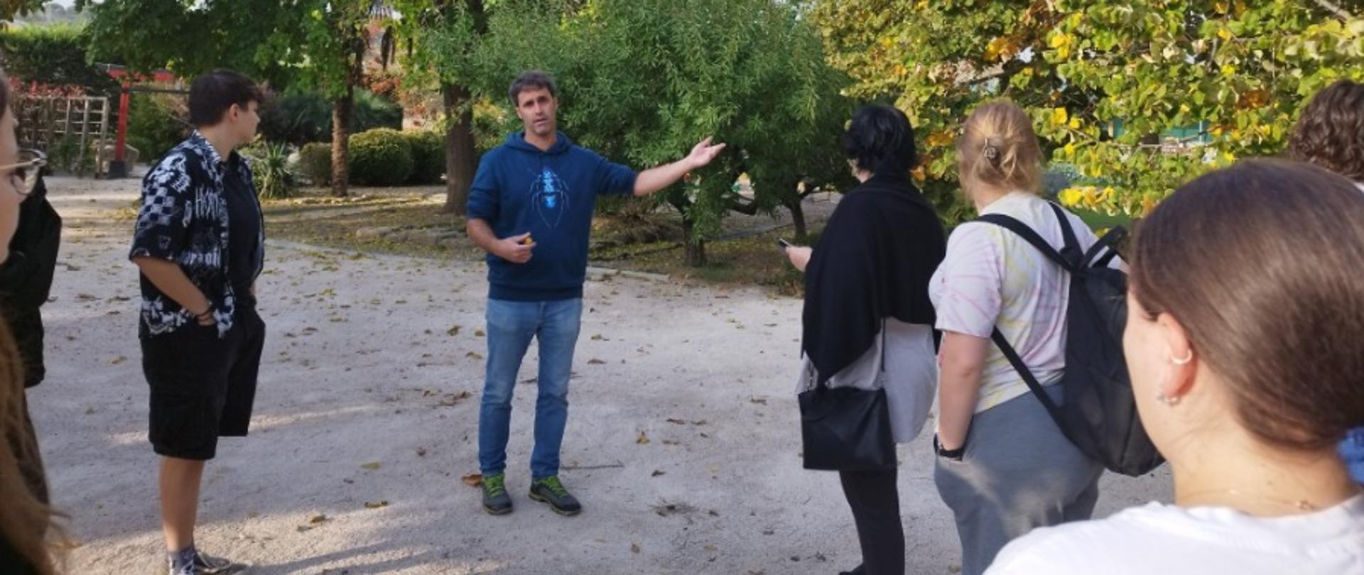 Grupa osób stoi na żwirowej ścieżce wśród drzew, a jedna osoba prowadzi omówienie roślin.