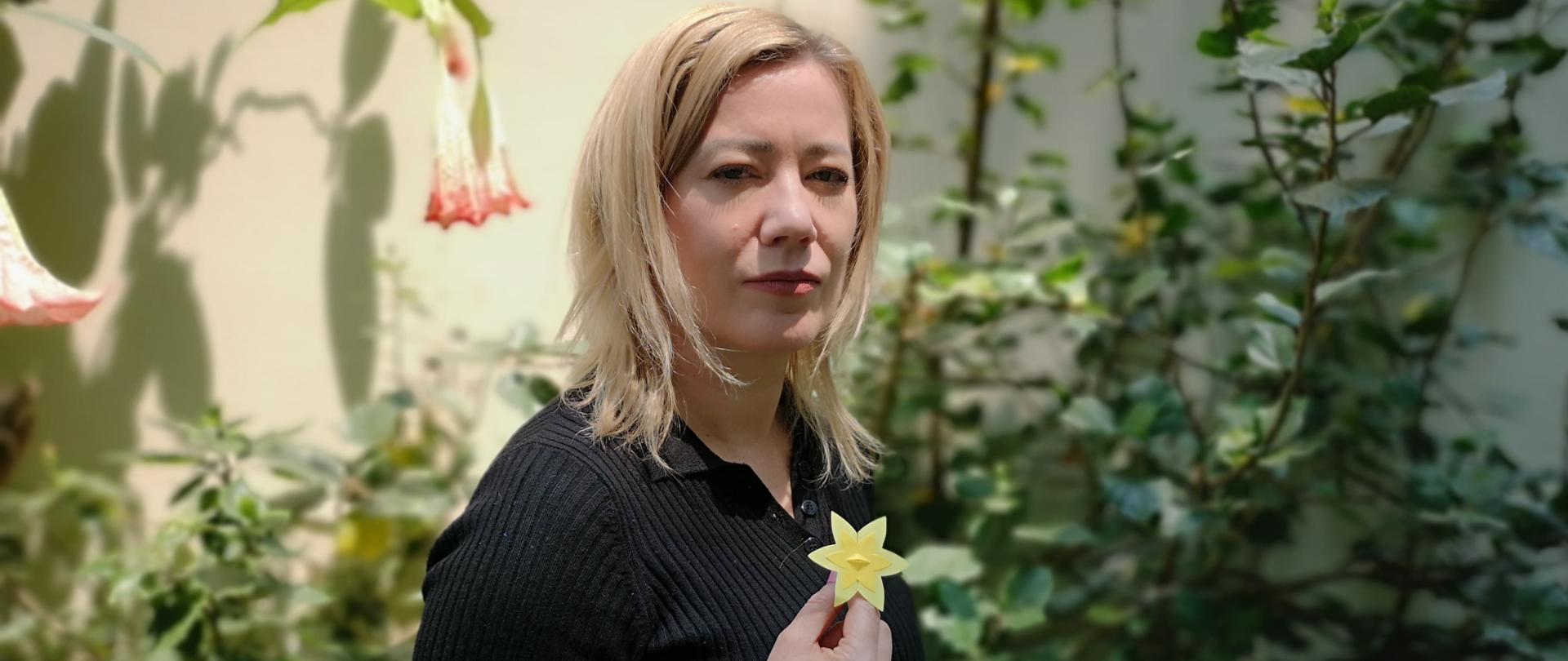 Embajadora Agnieszka Frydrychowicz-Tekieli, vestida de negro, mirando la camara. En el fondo jardin. En la mano tiene una flor amarilla - narciso, simbolo de levantamiento en el getto de Varsovia.