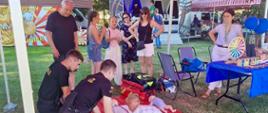 Pod czerwonym namiotem Komendy Powiatowej Państwowej Straży Pożarnej w Siemiatyczach strażacy prowadzą praktyczne zajęcia z pierwszej pomocy dla dzieci podczas Dni Siemiatycz. Uczestnicy ćwiczą resuscytację krążeniowo-oddechową na fantomie. W tle stoją mieszkańcy obserwujący pokaz oraz stoisko Powiatowej Stacji Sanitarno-Epidemiologicznej z materiałami edukacyjnymi.