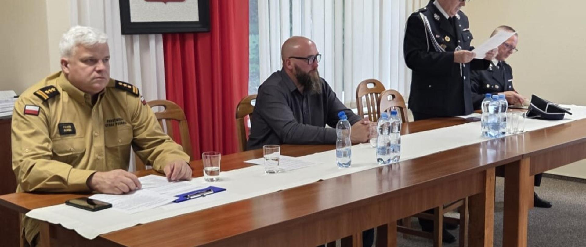 Osoby siedzące przy stole, mężczyzna w mundurze OSP czyta z kartki, w tle obraz z godłem RP.