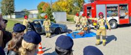 Na zdjęciu strażak tłumaczy zebranym uczniom szkoły ponadgimnazjalnej kolejność działań przy wypadku drogowym. Po prawej stronie widać pojazd pożarniczy oraz wrak samochodu osobowego. Strażacy w umundurowaniu specjalnym przygotowują się do przedstawienia technik cięcia poszczególnych elementów karoserii pojazdu.