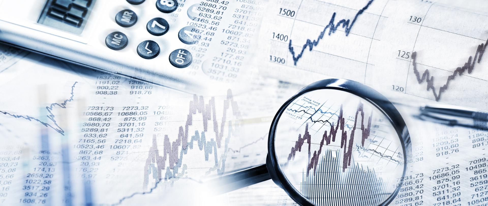 Na obrazie lupa, wykresy giełdowe oraz kalkulator i długopis