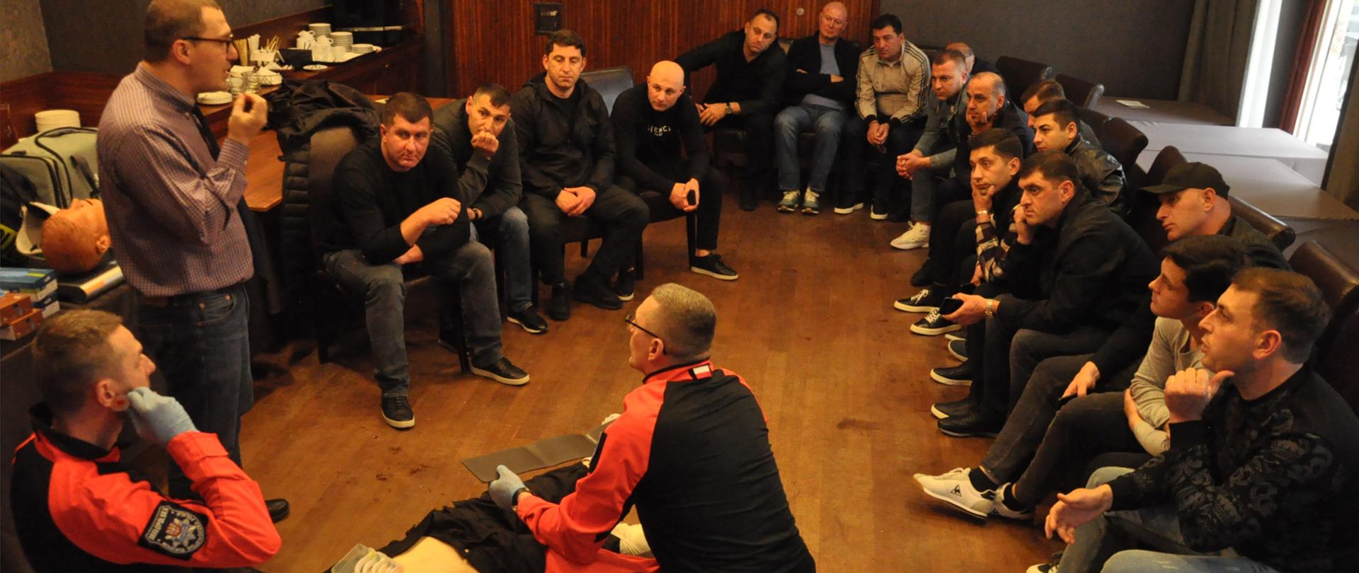 Grupa mężczyzn siedzi wokół leżącego na podłodze fantomu i słucha instruktora oraz dwóch ratowników demonstrujących udzielanie pierwszej pomocy 