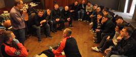 Grupa mężczyzn siedzi wokół leżącego na podłodze fantomu i słucha instruktora oraz dwóch ratowników demonstrujących udzielanie pierwszej pomocy
