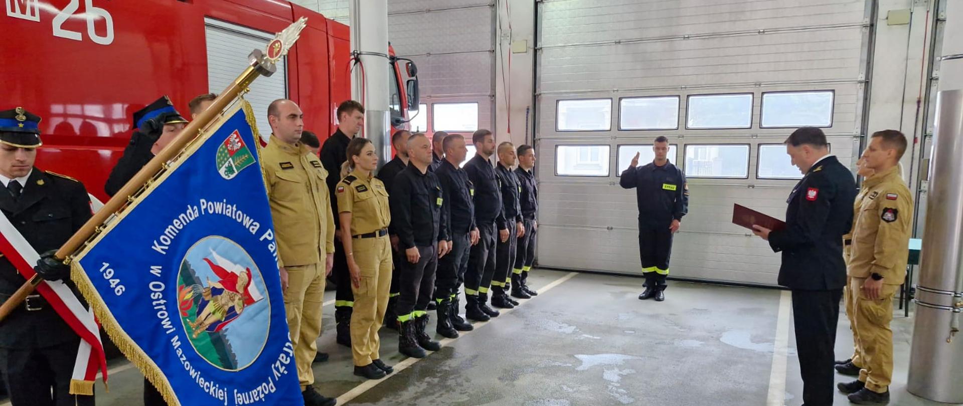 To zdjęcie, również zrobione wewnątrz remizy, przedstawia moment wręczania aktu ślubowania. Dwóch strażaków stoi przodem do siebie, uściągając sobie dłonie w uścisku, symbolizującym przyjęcie nowego strażaka do służby. Strażak w czarnym, polowym mundurze trzyma w rękach czerwone dokumenty. Starszy stopniem dowódca w granatowym, galowym mundurze, z licznymi odznaczeniami na piersi, przekazuje mu dokumenty. W tle stoi grupa innych strażaków, ubranych w różne mundury, będących świadkami tej ceremonii. W głębi widać również czerwony wóz strażacki.