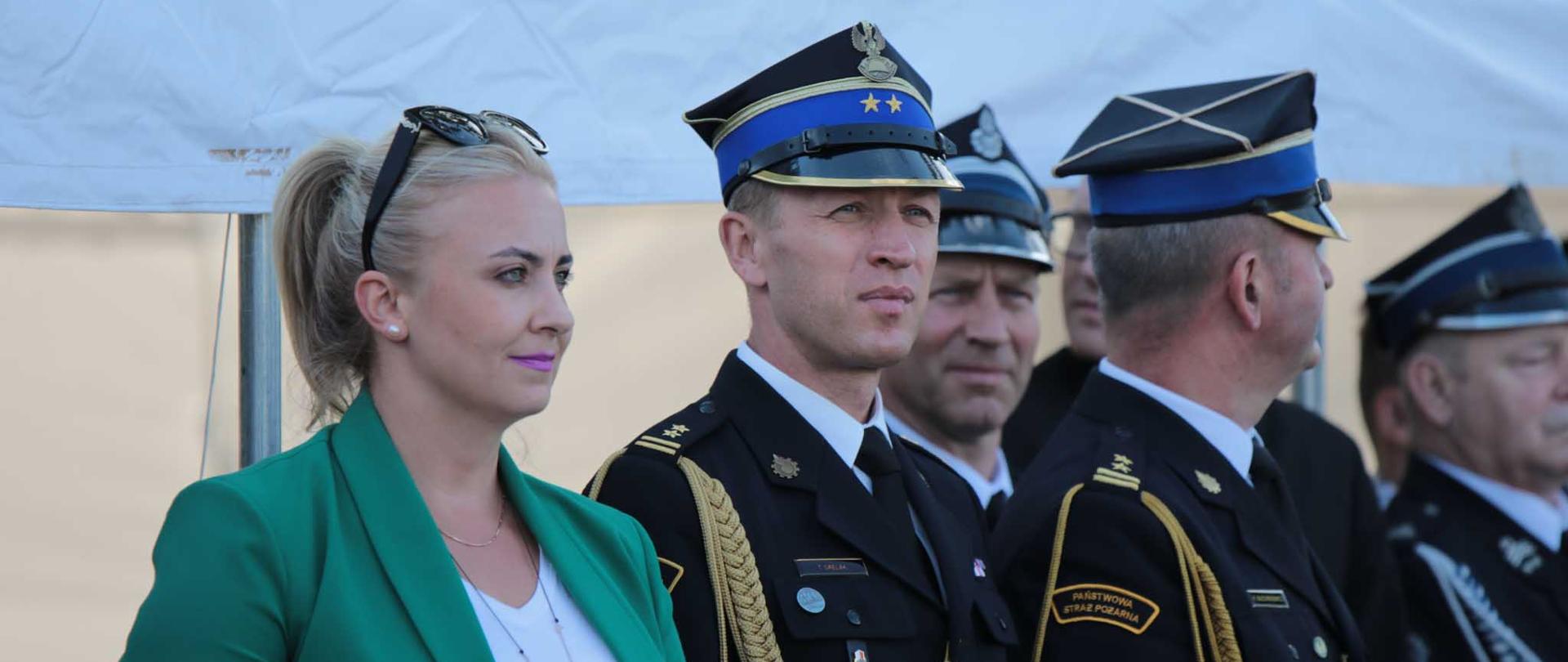 grupa ludzi stojaca po białym namiotem, na pierwzym planie kobieta w zielonej marynarce i białej koszuli, obok stoi strażak w rogatywce i ciemnym mundurze, w tle strażacy