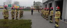 Zdjęcie przedstawia uroczystą zmianę służby podczas obchodów Święta Flagi. Dwa szeregi strażaków w umundurowaniu specjalnym koloru piaskowego i czerwonymi hełmami stoją w odstępach naprzeciwko siebie. Pośrodku szeregów st. kpt. Mariusz Wenta z-ca dowódcy JRG w umundurowaniu wyjściowym. Strażacy na twarzach mają założone maseczki ochronne. Po prawej stronie widoczny budynek strażnicy