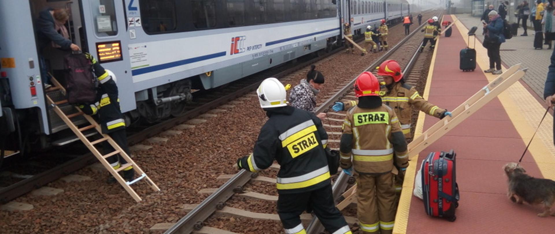 Na zdjęciu widać po lewej stronie pociąg relacji Przemyśl – Wrocław do jego wejścia przystawiona jest drabina po której schodzi kobieta za rękę trzyma ja strażak ubrany w czarny mundur z seledynowymi paskami i białym hełmie. po prawej stronie dostawiona jest drabinka do wejścia na peron przy niej stoi dwóch strażaków w piaskowym mundurze i czerwonych hełmach i jeden w czarnym mundurze i białym hełmach. Po prawej stronie na peronie stoją ludzie. Przy drabince na peronie stoi czerwony bagaż oraz mężczyzna z mały pies
