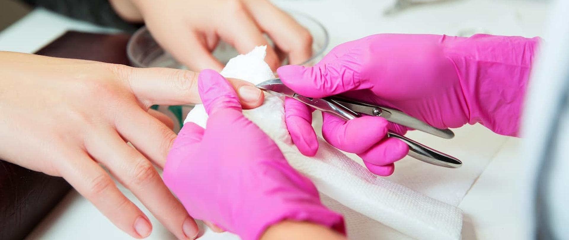 Zdjęcie przedstawia dłonie kosmetyczki w różowych, gumowych rękawiczkach dokonujące manicure klientce.