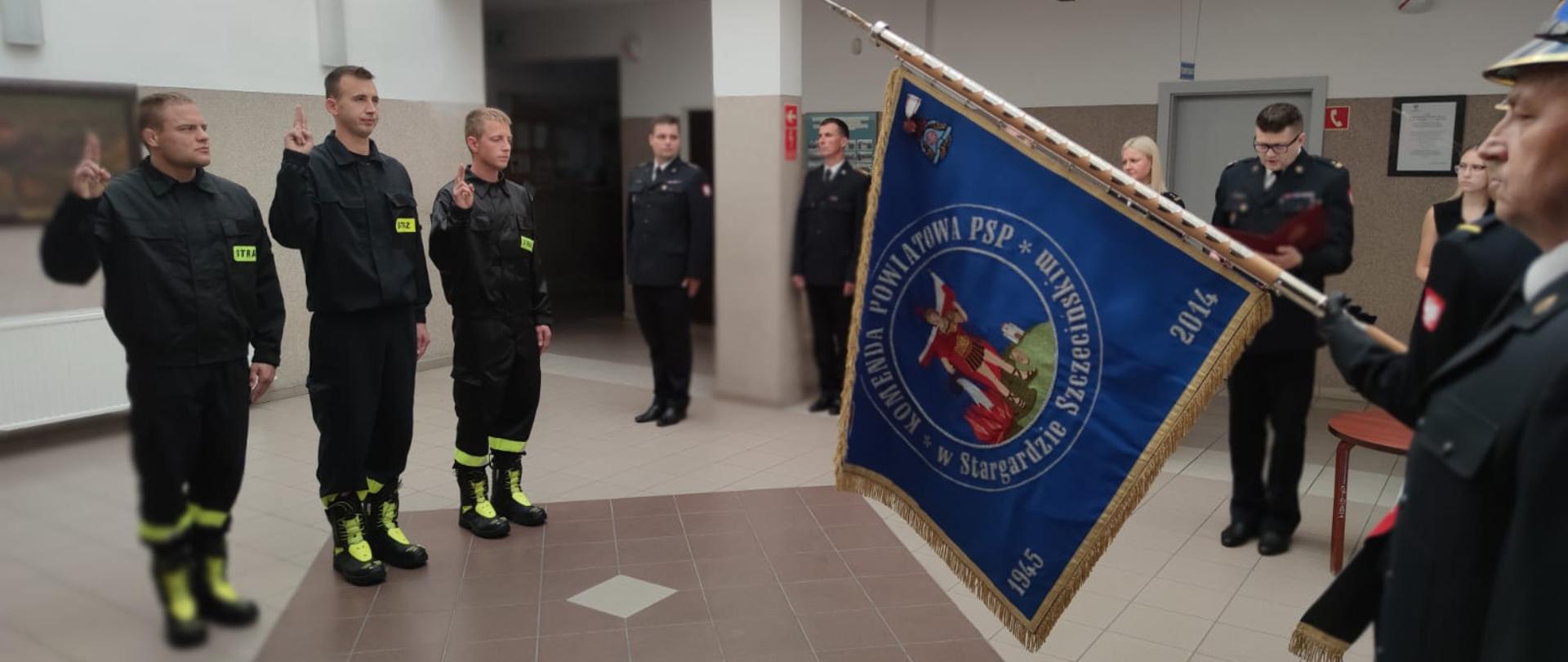 Przed sztandarem Komendy Powiatowej PSP w Stargardzie Szczecińskim stoi trzech strażaków trzymają prawe ręce przedramieniem pionowo do góry ze złączonymi i wyprostowanymi wskazującymi i środkowymi palcami.