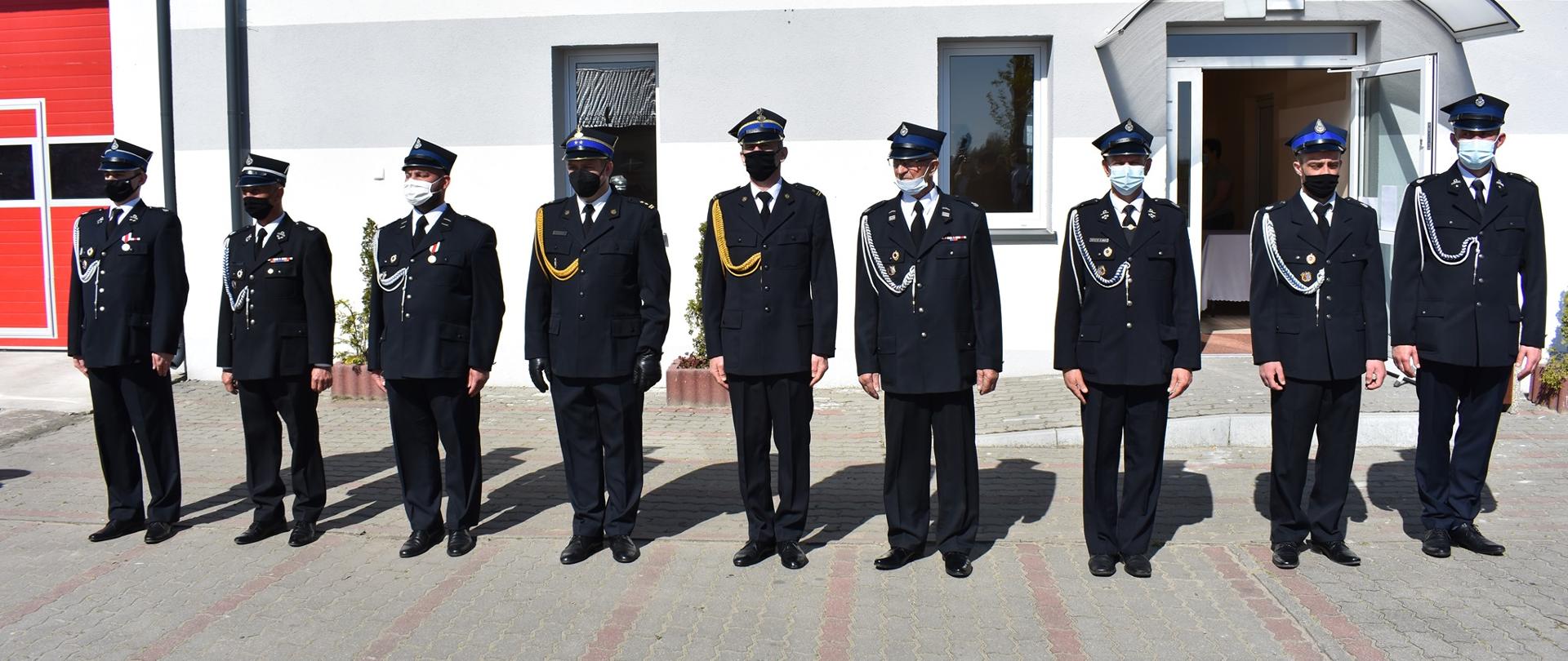 Zdjęcie przedstawia strażaków PSP oraz OSP stojących w szeregu. Wszyscy ubrani są w mundury wyjściowe oraz mają założone na twarzy maseczki chirurgiczne. Strażacy stoją w pozycji na baczność i czekają na odznaczenia. W tle zdjęcia znajduje się budynek remizy OSP.