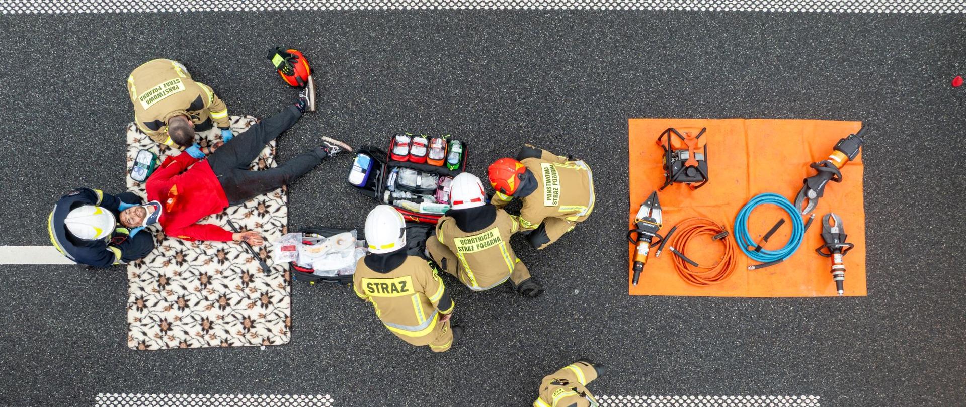 Kadr z góry przedstawiający strażaków udzielających pomocy poszkodowanemu na kocu oraz rozłożony obok sprzęt medyczny i hydrauliczny na pomarańczowej macie. W rogu widoczny jest przód wozu strażackiego.