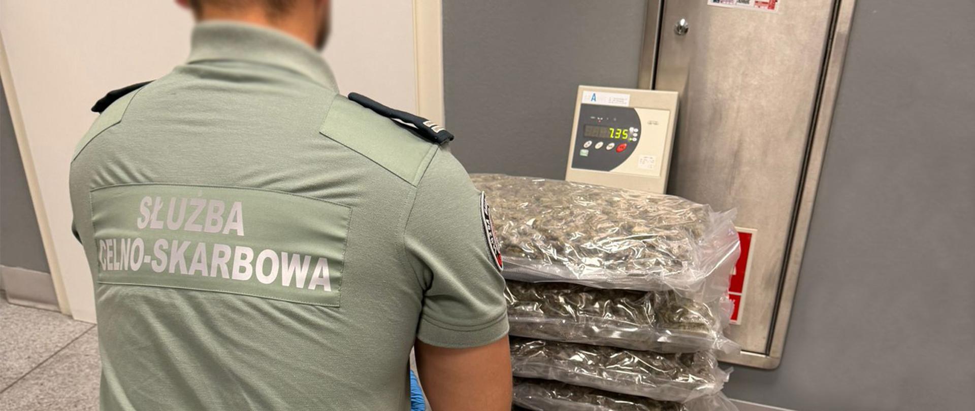 Funkcjonariusz w koszulce z napisem „Służba Celno‑Skarbowa” przy wadze, na której ułożono kilka dużych pakunków.