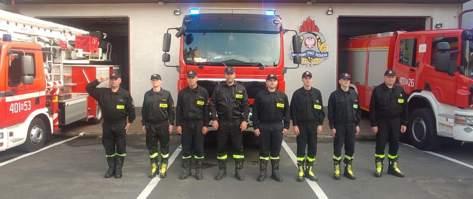 Na zdjęciu widać strażaków z KP PSP w Ustrzykach Dolnych w ubraniach koszarowych stojących na baczność. Pierwszy od lewej czyli dowódca oddaje honor. Za nimi stoją wozy bojowe z włączonymi sygnałami oraz widać budynek Komendy. 
