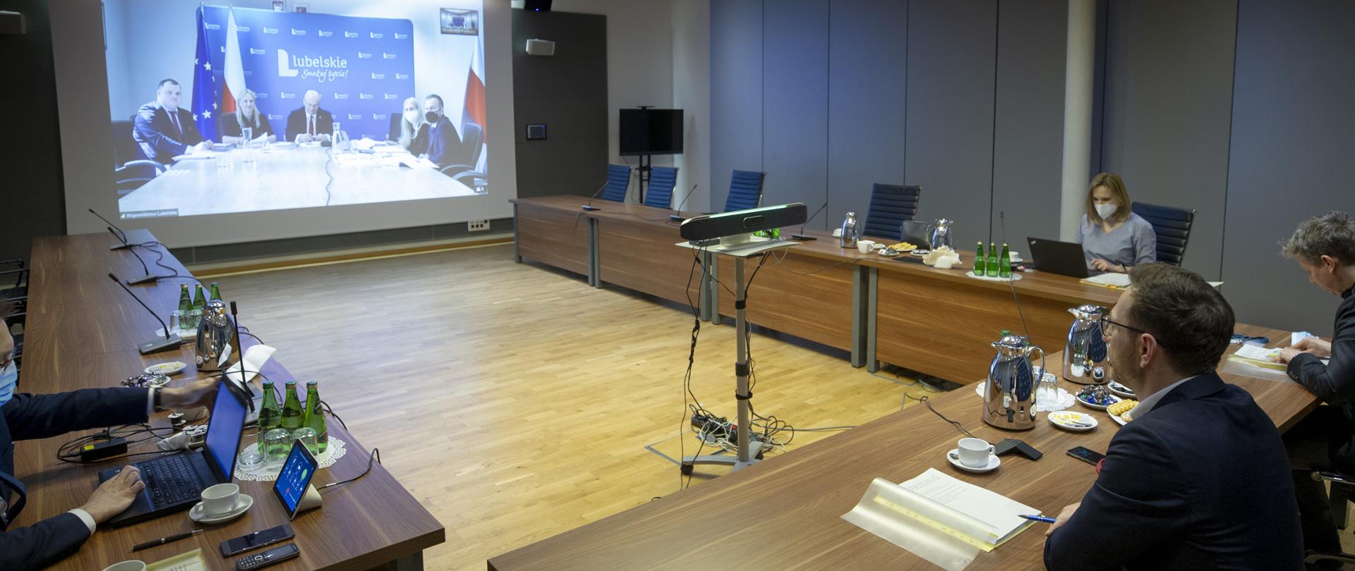 Na sali konferencyjnej przy stołach ustawionych w podkowę siedzą osoby. Przed nimi duży monitor.